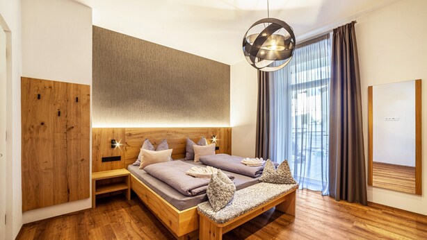 A modern bedroom with a wooden bed, a bench, a large window with curtains, and a unique spherical light fixture.