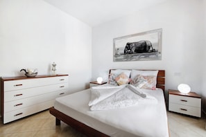 Un dormitorio con cama, mesitas de noche, cómoda, un cuadro en la pared y una mesilla con lámpara.