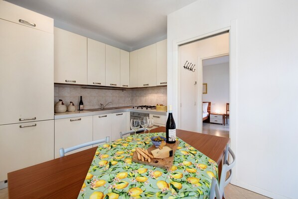 A modern kitchen with white cabinets, a dining table with a fruit-patterned tablecloth, and a bottle of wine.