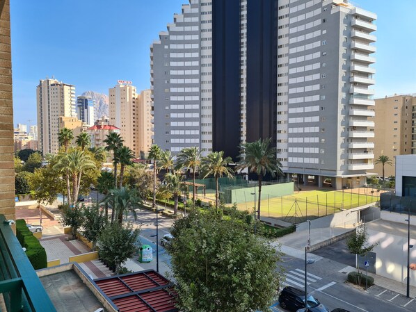 A high-rise building with multiple balconies, a sports field, and surrounding palm trees.