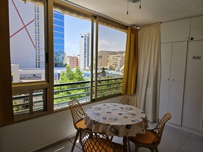 A room with a balcony, a table with a patterned tablecloth, and a view of buildings and greenery.