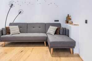 A grey sectional sofa with wooden legs, a wooden floor, and a wall-mounted shelf with decorative items.