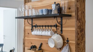 Eine Küche mit einer Holzwand, einem schwarzen Regal mit Gläsern und Tassen, einem hängenden Gestell mit weißen Tassen und einer Holzkiste mit Besteck.