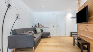 Ein modernes Wohnzimmer mit grauem Sofa, Holzboden, einem an der Wand befestigten Fernseher und einer Tür im Hintergrund.
