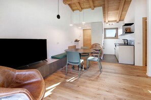 A modern living space with a flat-screen TV, a dining area with a table and chairs, a kitchen with a refrigerator, and a wooden ceiling.