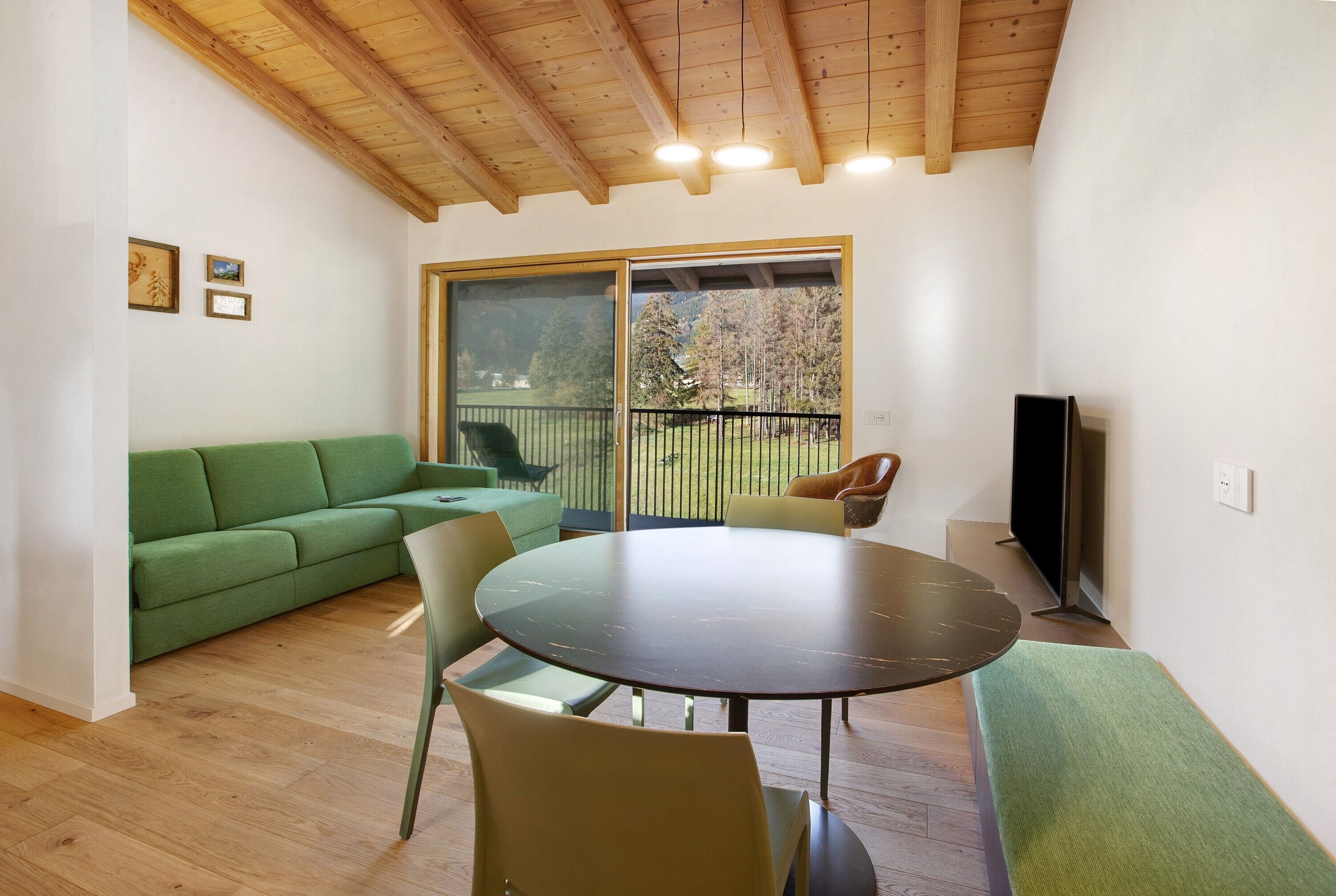 Een kamer met een groene bank, een ronde tafel, stoelen, een televisie en een groot raam met uitzicht op bomen.