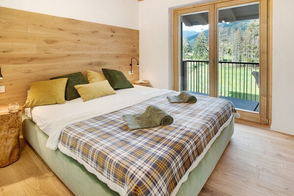 Un dormitorio con cabecera de madera, cama con ropa de cama escocesa, mesita de noche con lámpara y vistas a un balcón con barandilla.