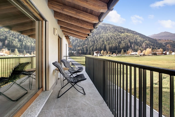 A balcony with two chairs, a glass table, and a view of a forested mountain.