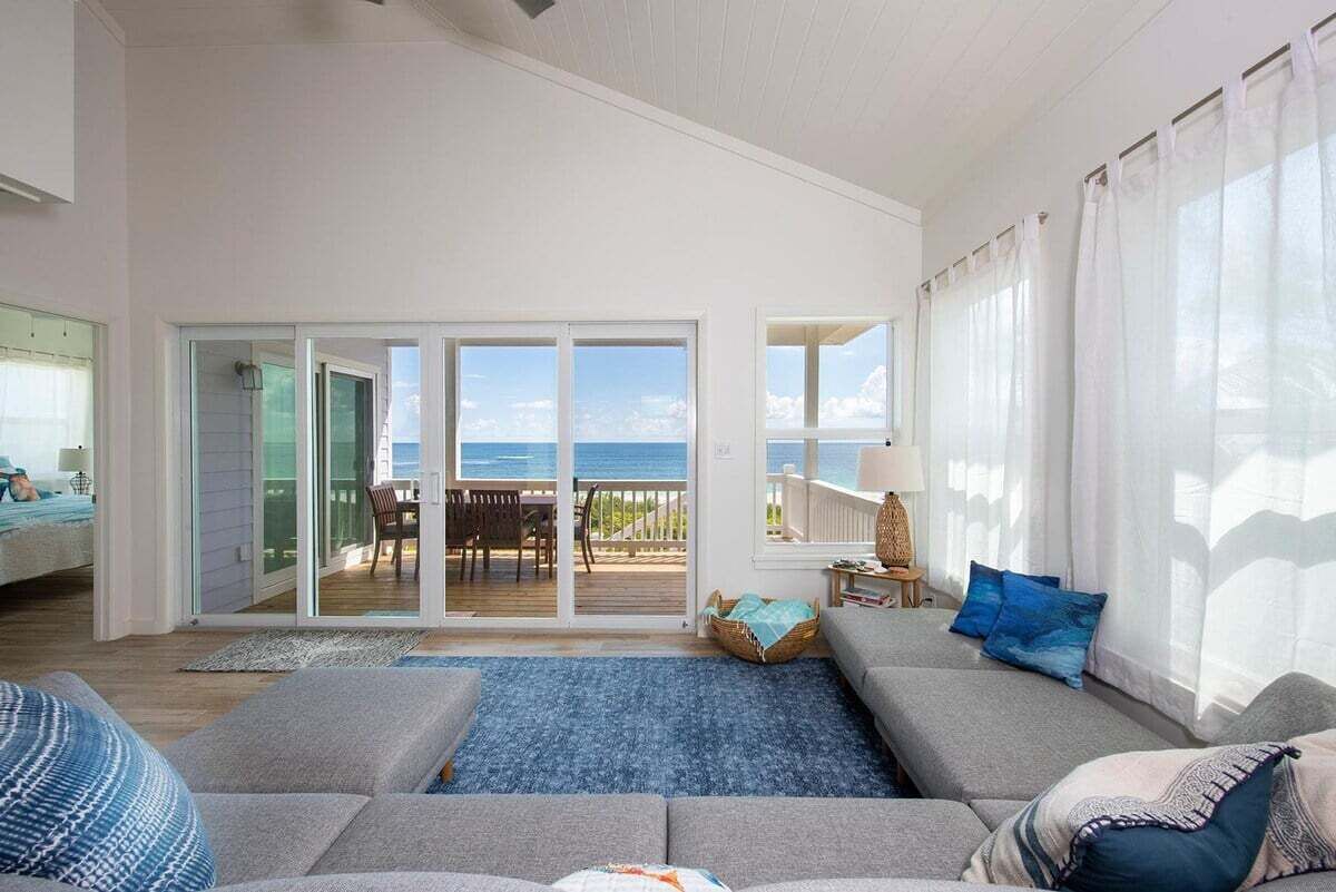 A spacious living room with a large grey sofa, a blue rug, and a view of the ocean through a sliding glass door.