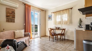 A living room with a sofa, a dining table with chairs, a kitchen area with a refrigerator, and a sliding glass door leading to a balcony.