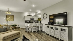 Una sala moderna con un sofá, una mesa de centro y un televisor. Hay una zona de cocina con armarios blancos y una mesa de comedor.
