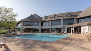 Un área de piscina con sillones y sombrillas, un edificio de piedra con grandes ventanales y un cielo despejado.