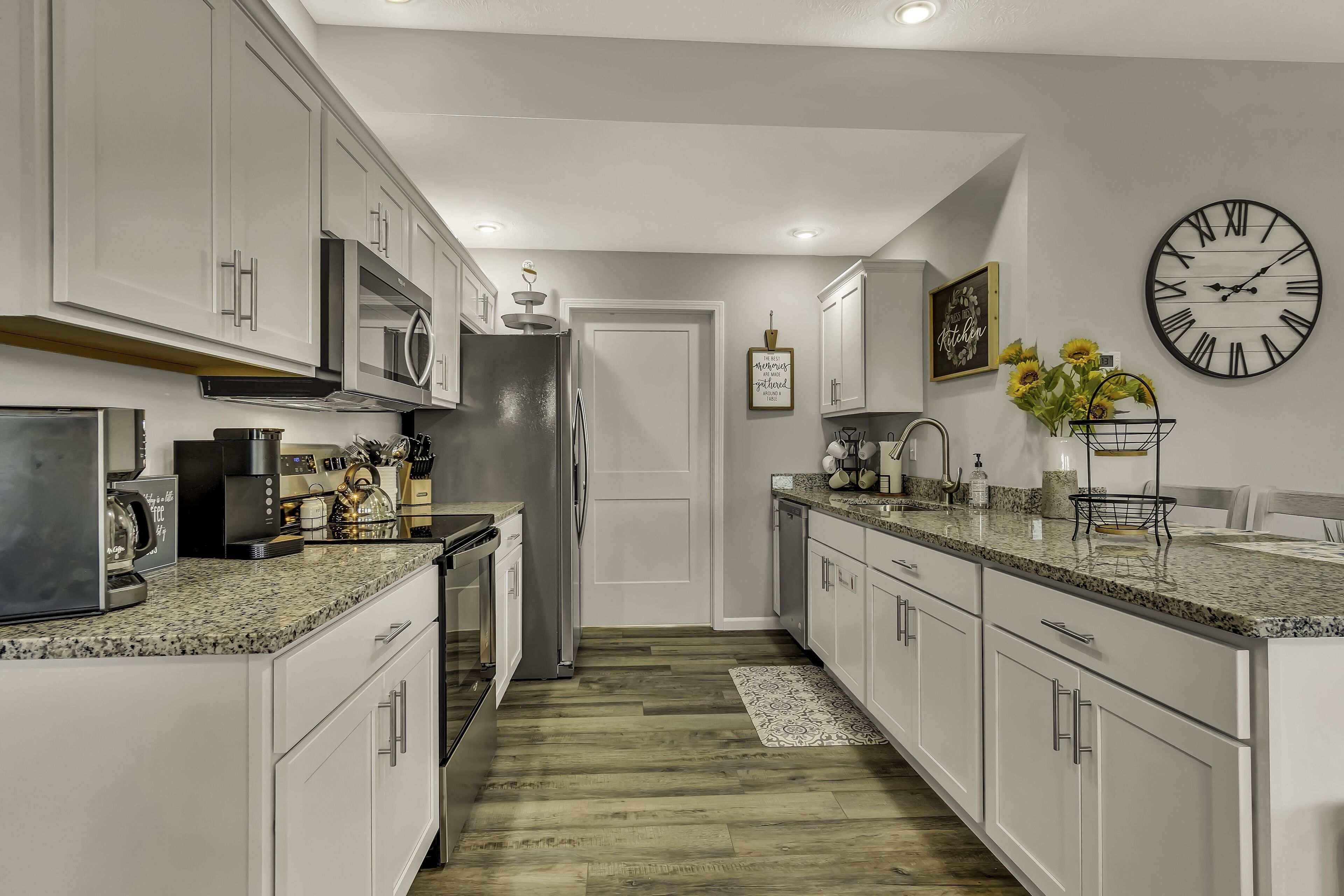 A modern kitchen with white cabinets, granite countertops, a stainless steel refrigerator, and a large wall clock.