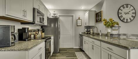 A modern kitchen with white cabinets, granite countertops, a stainless steel refrigerator, and a large wall clock.