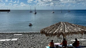 En strand med en stråtagsparasol, en klippefyldt kystlinje og sejlbåde i vandet.