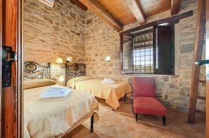 Une pièce avec deux lits, une chaise rouge, des murs en pierre, une fenêtre avec des barreaux et un plafond en bois.