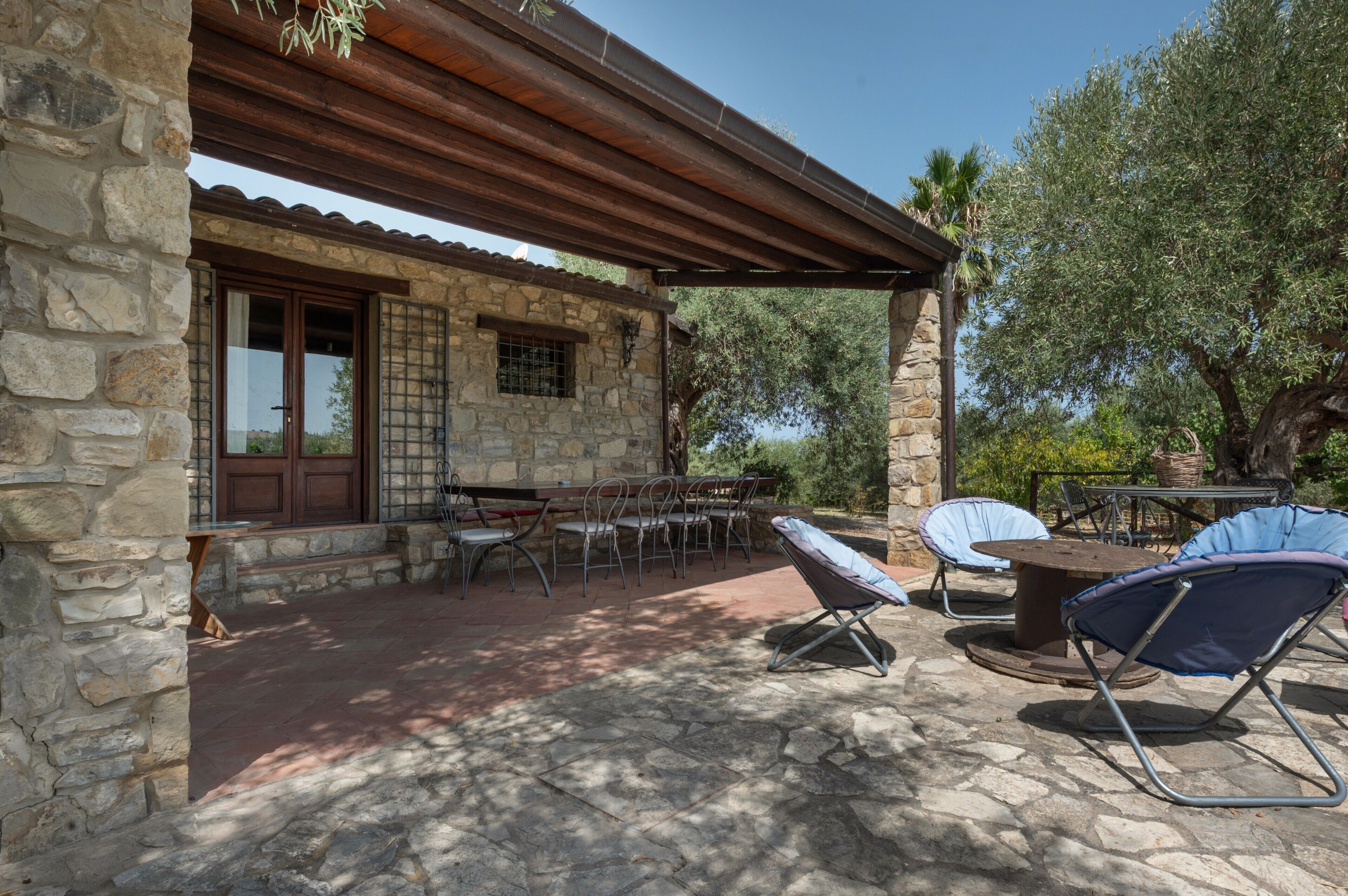 A stone building with a covered patio, outdoor seating, and olive trees.