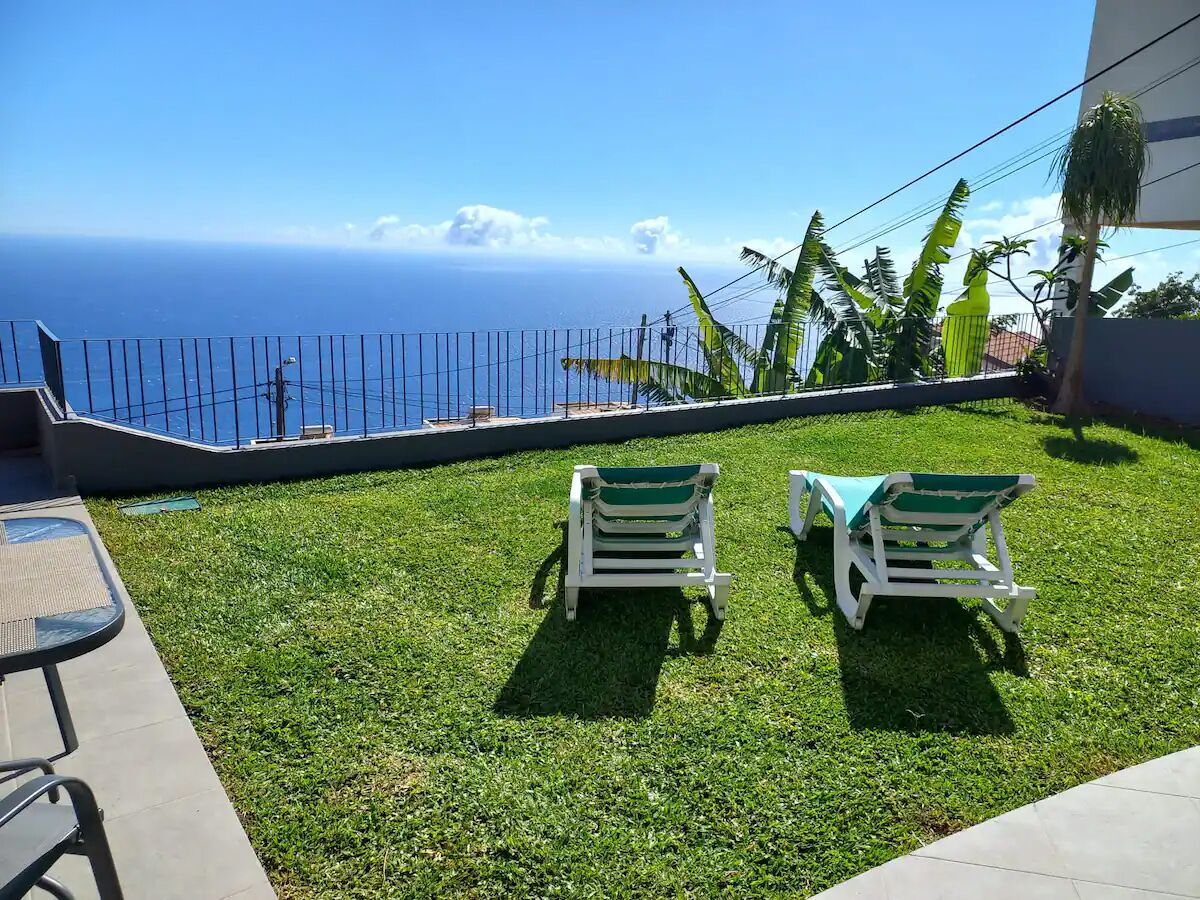 A grassy area with two white lawn chairs and a table, overlooking a body of water.