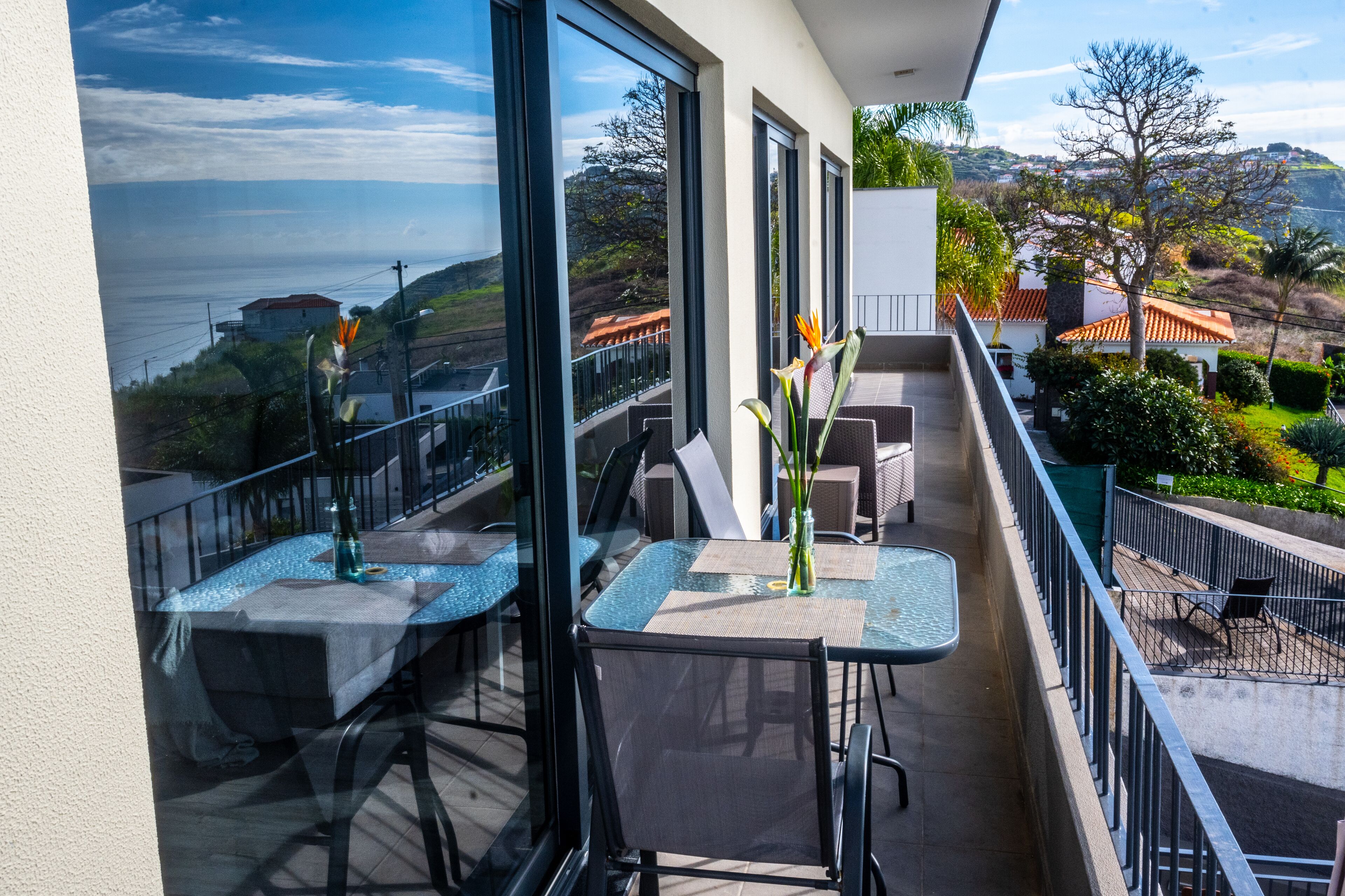 A balcony with tables and chairs, overlooking a scenic view of the sea and hills.