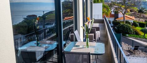 A balcony with tables and chairs, overlooking a scenic view of the sea and hills.