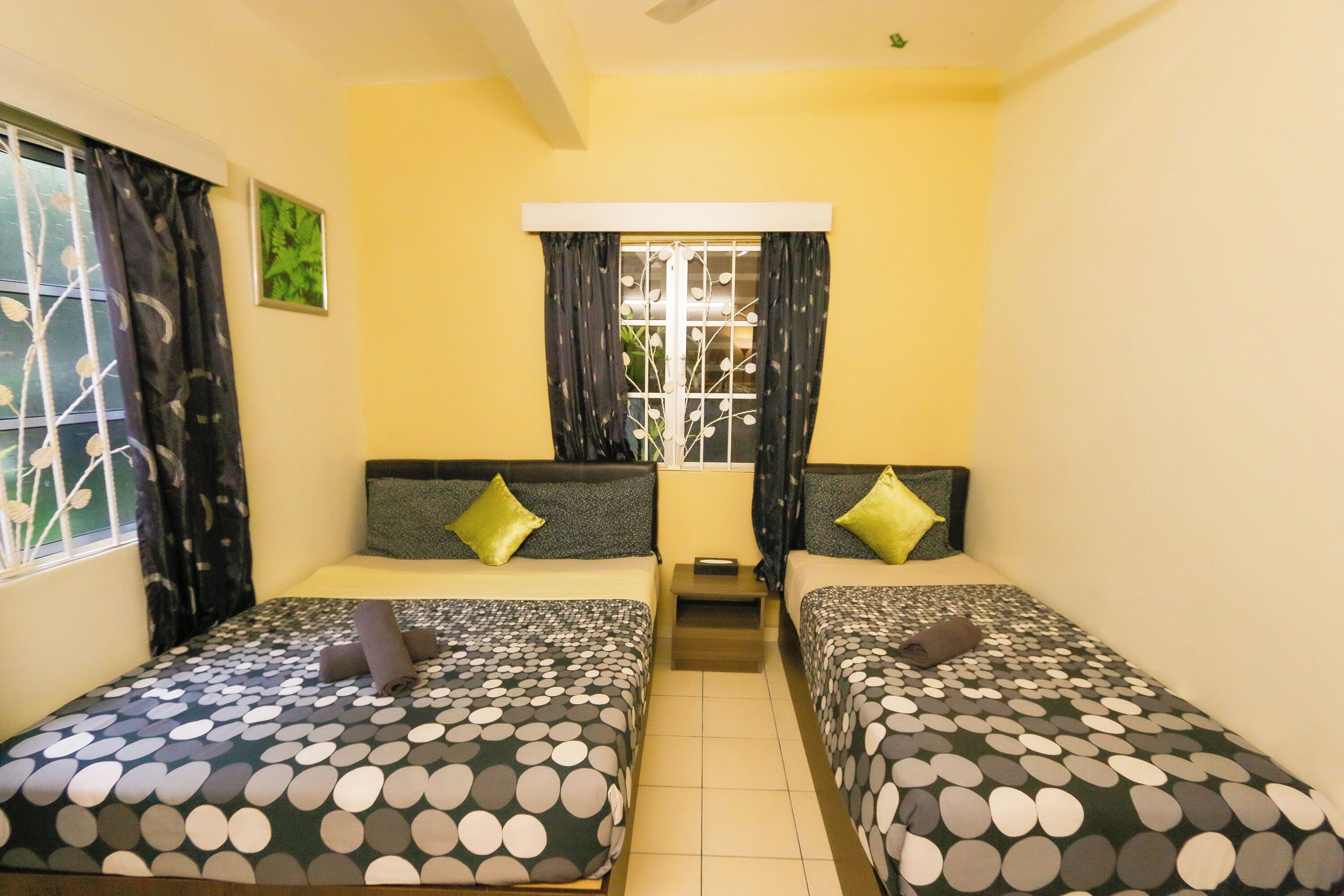 A room with two single beds, each with a patterned bedspread and pillows, a small bedside table, and a window with curtains.