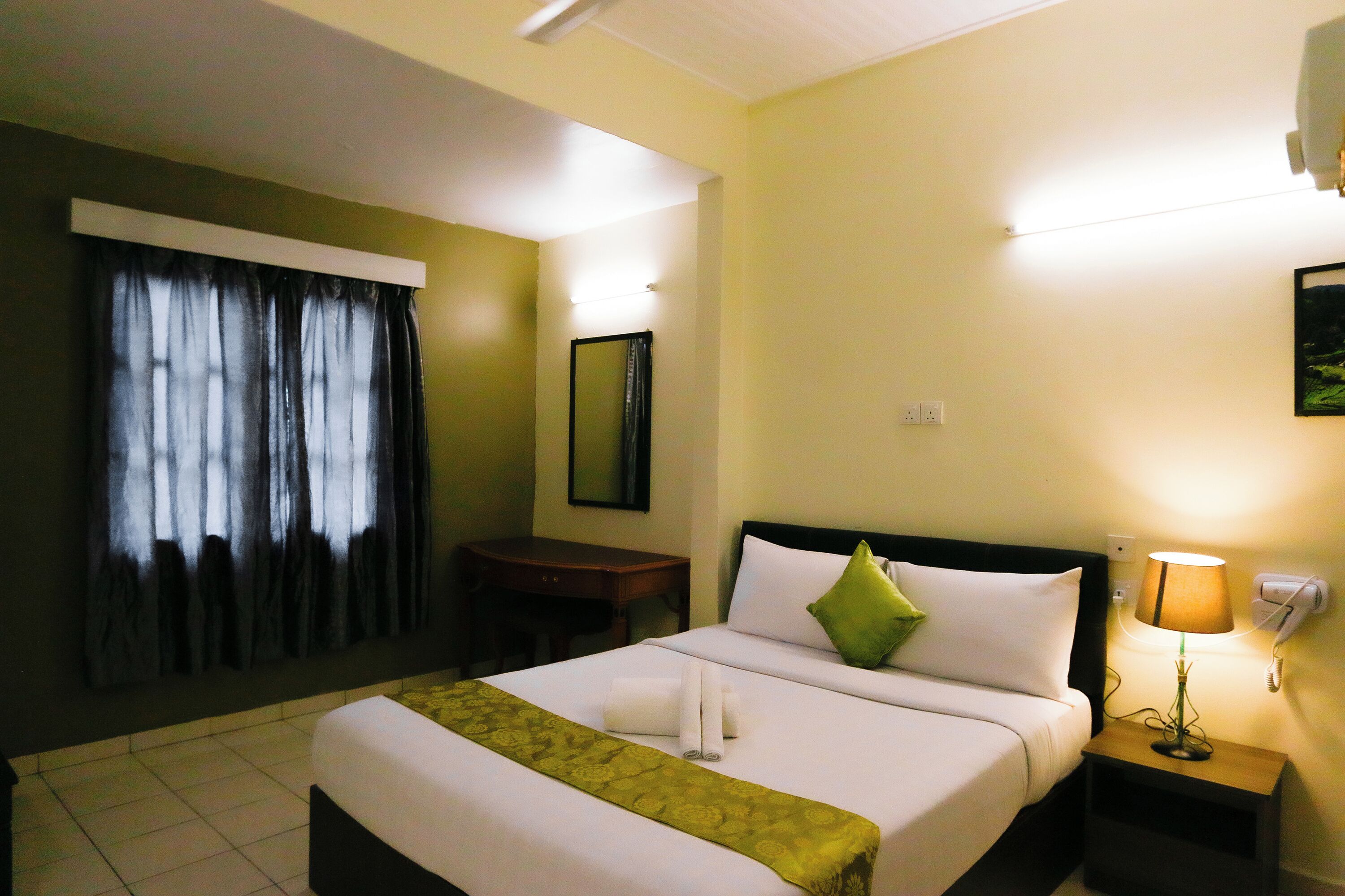 A hotel room with a large bed, a bedside table with a lamp, a desk, and a window with curtains.