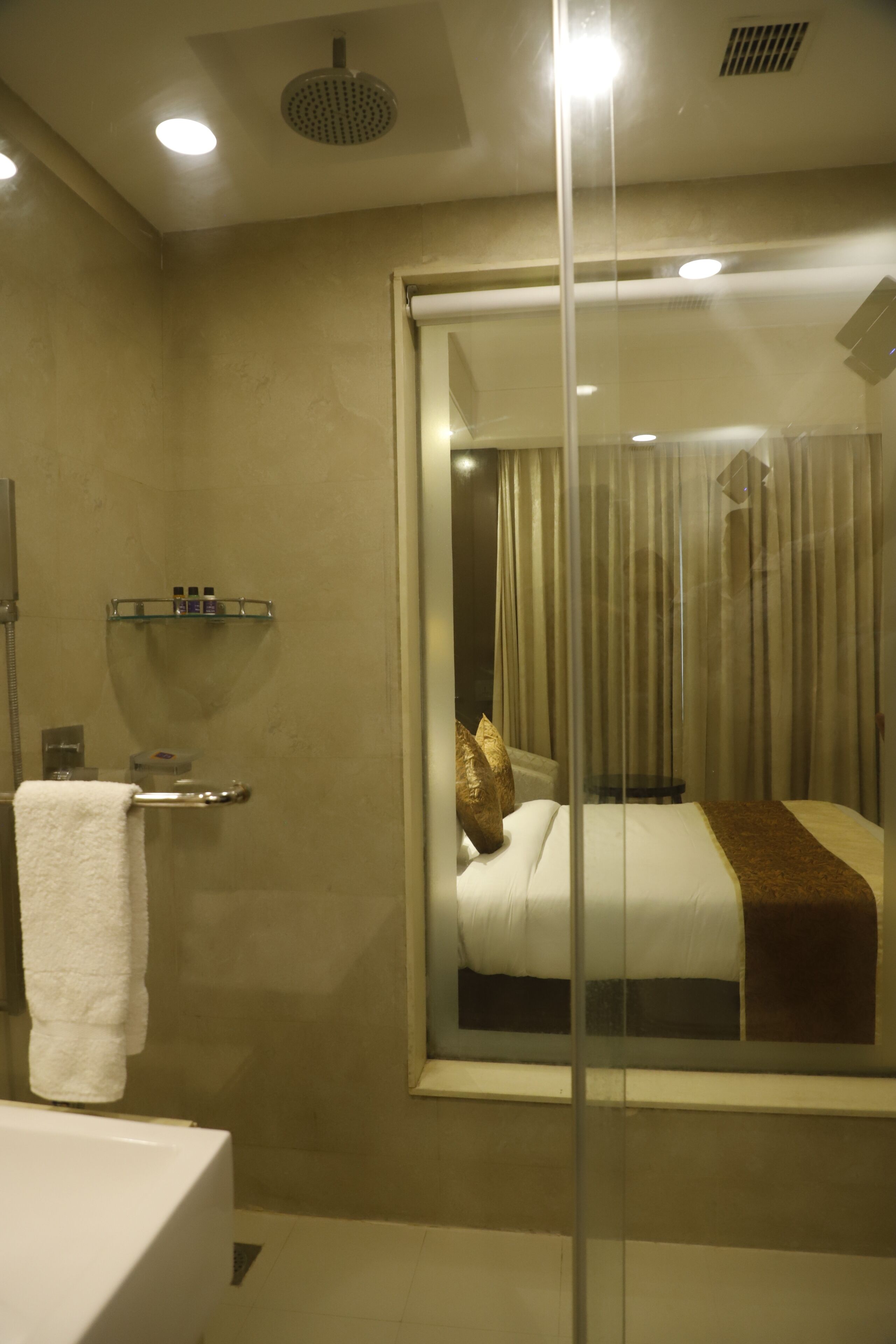A hotel room with a glass partition, a shower, a towel rack, and a bed with a brown throw blanket.