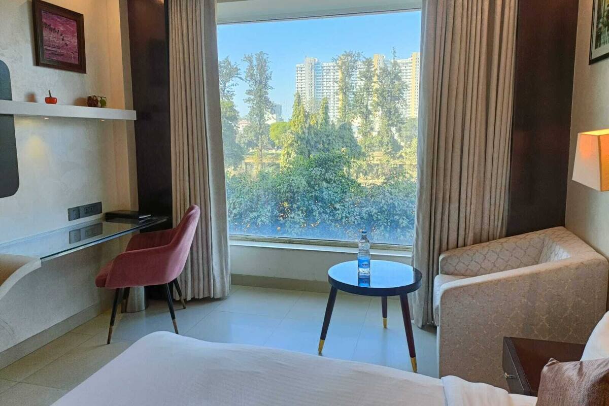 Habitación de hotel con cama, escritorio, silla, una mesita con una botella, y vistas a edificios y árboles a través de la ventana.