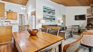 A spacious living area with a wooden ceiling, a dining table with chairs, a fireplace, and a mounted television. - Whitefish Lake Condo, 14BU, hillside! (Whitefish)