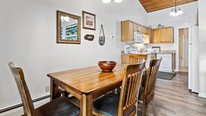 A dining area with a wooden table and chairs, a kitchen with wooden cabinets, and a white wall with framed pictures. - Whitefish Lake Condo, 14BU, hillside! (Whitefish)