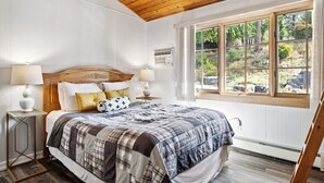 A bedroom with a wooden ceiling, a large bed with a plaid bedspread, two bedside lamps, and a window with a view of trees. - Whitefish Lake Condo, 14BU, hillside! (Whitefish)