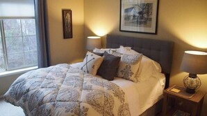 Een net opgemaakt bed met een bedsprei met dessin, twee bedlampen, een ingelijst beeld op de muur en een raam met gordijnen.