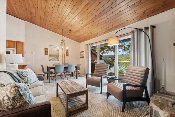 A living room with a wooden ceiling, a dining area, and a view of greenery outside.