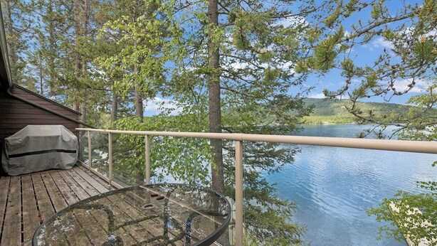 Um deck de madeira com mesa de vidro e guarda-corpo de metal, com vista para um lago.