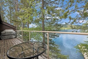A wooden deck with a glass table and a metal railing overlooking a lake.