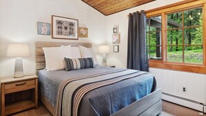 Um quarto com teto de madeira, uma cama com manta listrada, luminárias de cabeceira, quadros nas paredes e uma janela com vista para árvores.