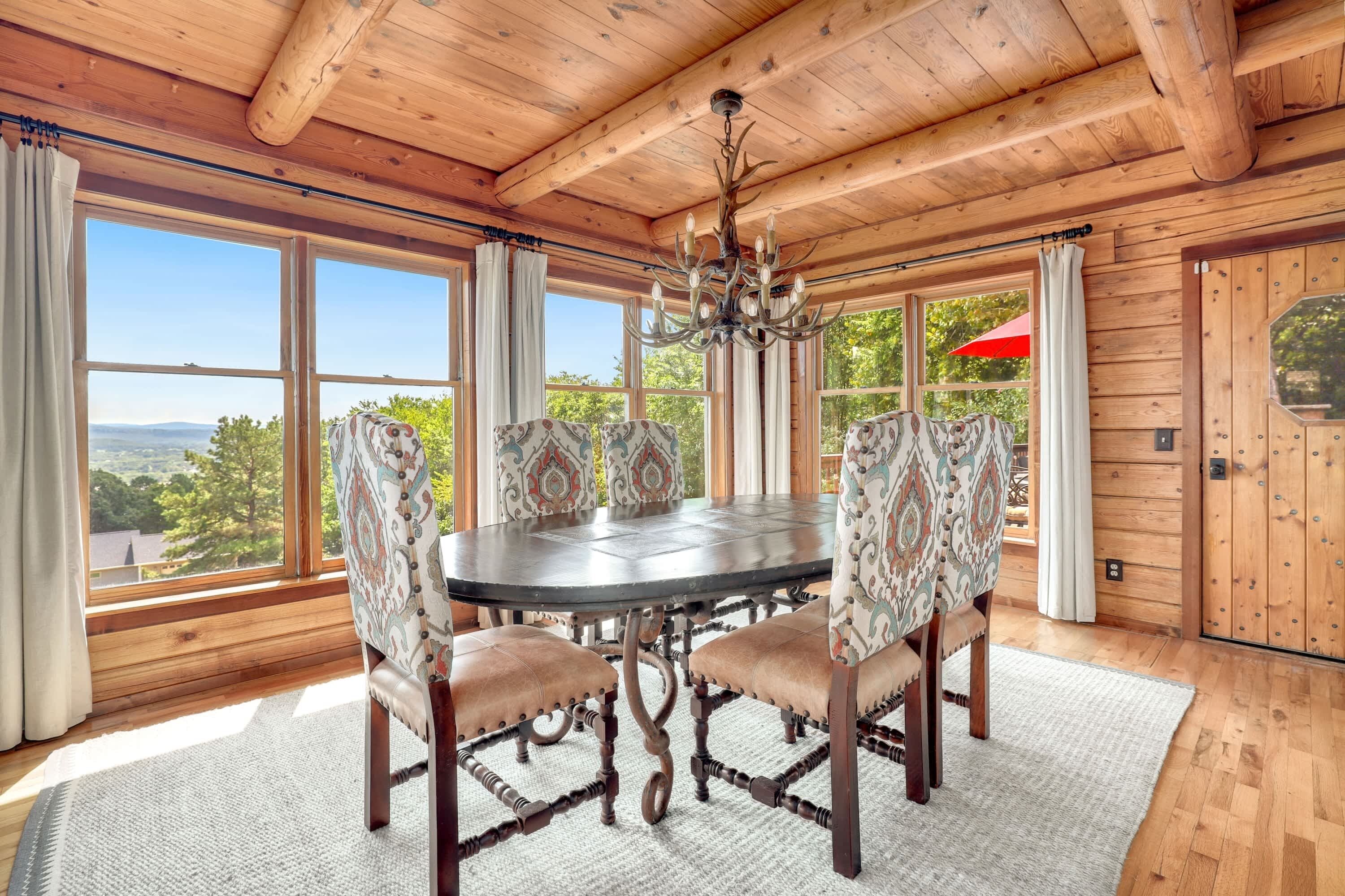 A wooden dining room with a round table and patterned chairs, large windows with curtains, and a chandelier.