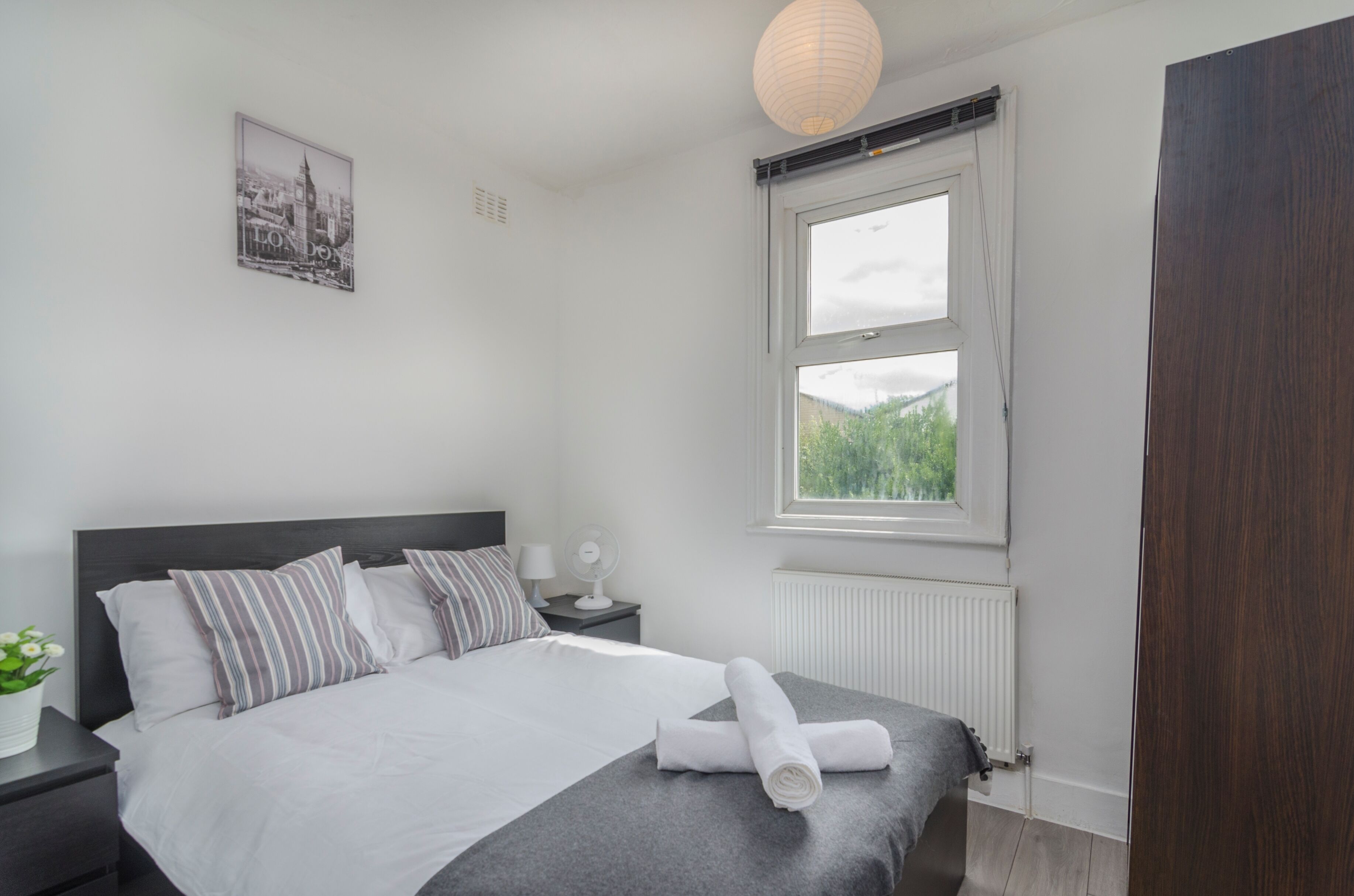 A modern bedroom with a large bed, a bedside table with a lamp, a window with a view of greenery, and a wall-mounted picture of a cityscape.