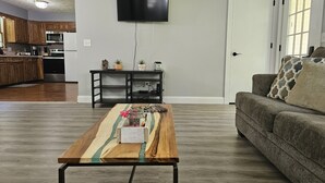 Un salon avec une table basse en bois, un canapé gris, un téléviseur à écran plat fixé au mur, et une cuisine en arrière-plan.
