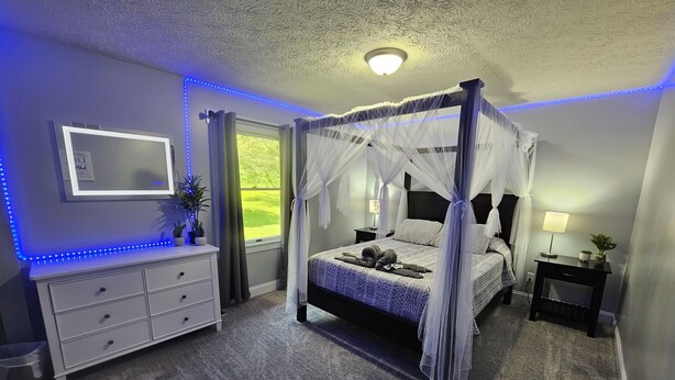 Une chambre à coucher avec un lit à baldaquin, une table de chevet avec une lampe, une commode et des bandes lumineuses à LED le long du plafond.