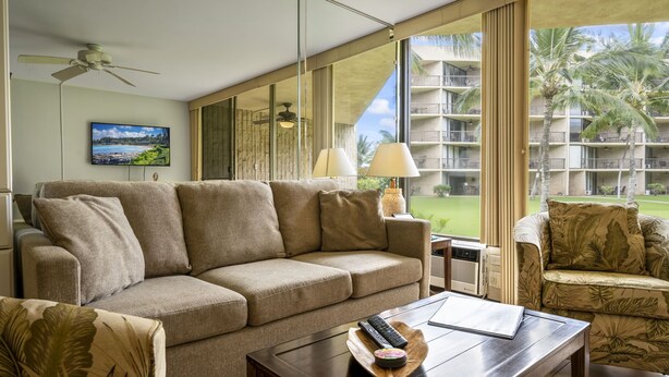 A modern living room with a beige sofa, a wooden coffee table, and a view of a hotel building and palm trees. - Steps to the ocean sunny Kihei, Maui Sunset A104 (Kihei)