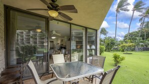 A patio with a table and chairs, a ceiling fan, and a view of a well-maintained lawn and palm trees. - Steps to the ocean sunny Kihei, Maui Sunset A104 (Kihei)