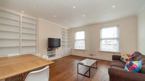 Ein modernes Wohnzimmer mit einem Holzesstisch, weißen Stühlen, einem braunen Sofa mit bunten Kissen, einem schwarzen Fernseher auf einem Schrank und integrierten Regalen.
