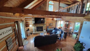 A log cabin interior with a fireplace, a black leather sofa, wooden floors, and a TV mounted on the wall. - 20% OFF Lake View Retreat- Lake & Mountain View, Hot Tub, Fire Pit, Porch Swing (Dandridge)