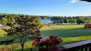 A view from a covered porch overlooking a grassy area with a lake and houses in the distance. - 20% OFF Lake View Retreat- Lake & Mountain View, Hot Tub, Fire Pit, Porch Swing (Dandridge)