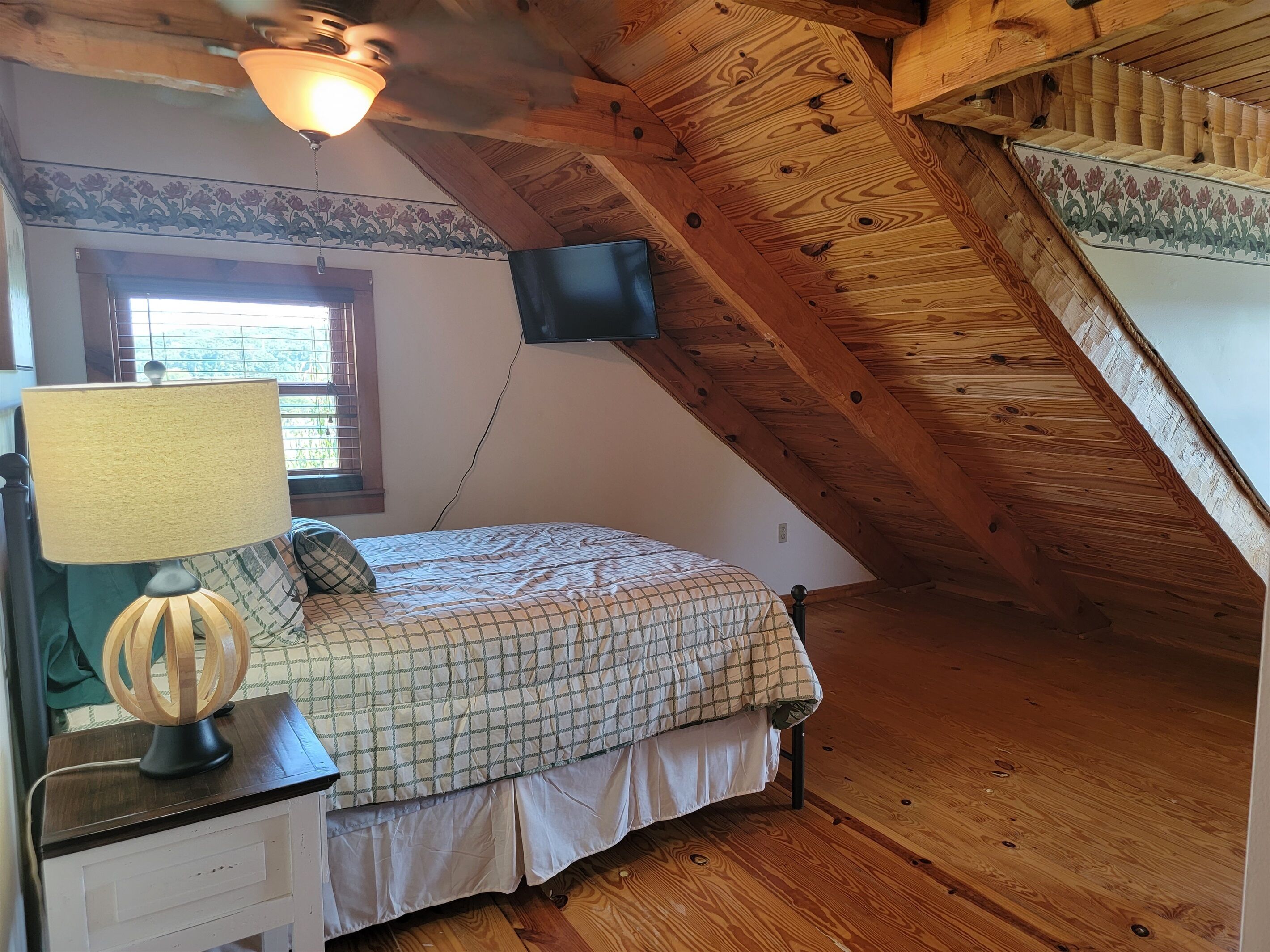 Ein Schlafzimmer mit einem Bett, einem Nachttisch, einer Lampe, einem Fenster und einem an der Wand befestigten Fernseher.