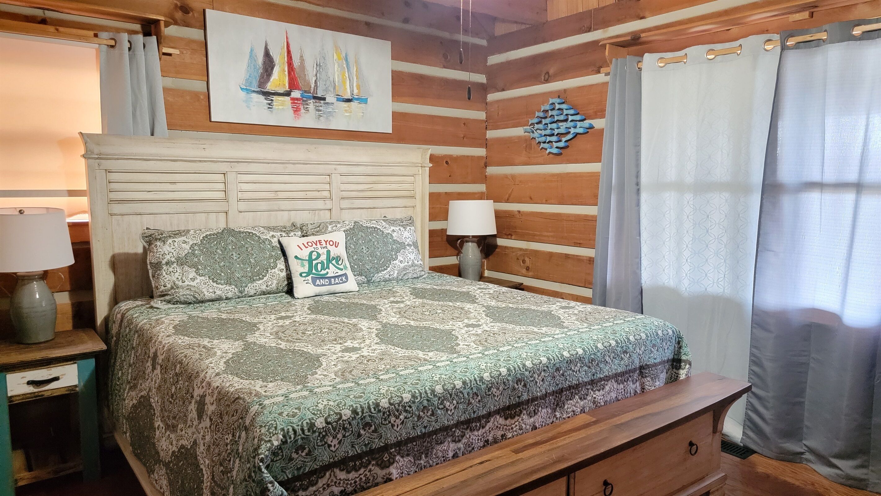 A wooden cabin-style bedroom with a bed, two pillows, a nightstand, a lamp, a painting of sailboats, and a wall decoration.