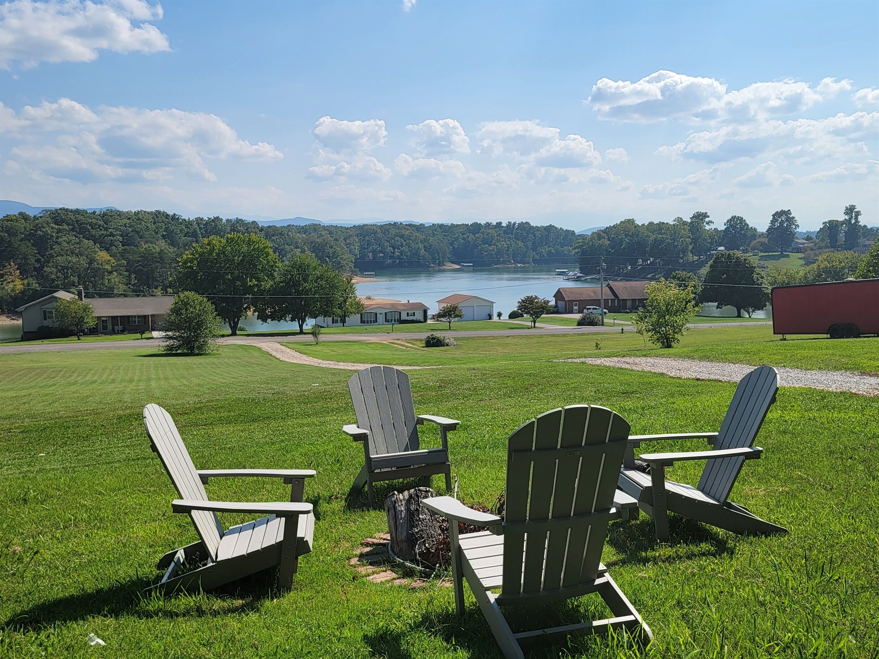 A grassy outdoor area with Adirondack chairs and a view of a lake and buildings in the distance.
