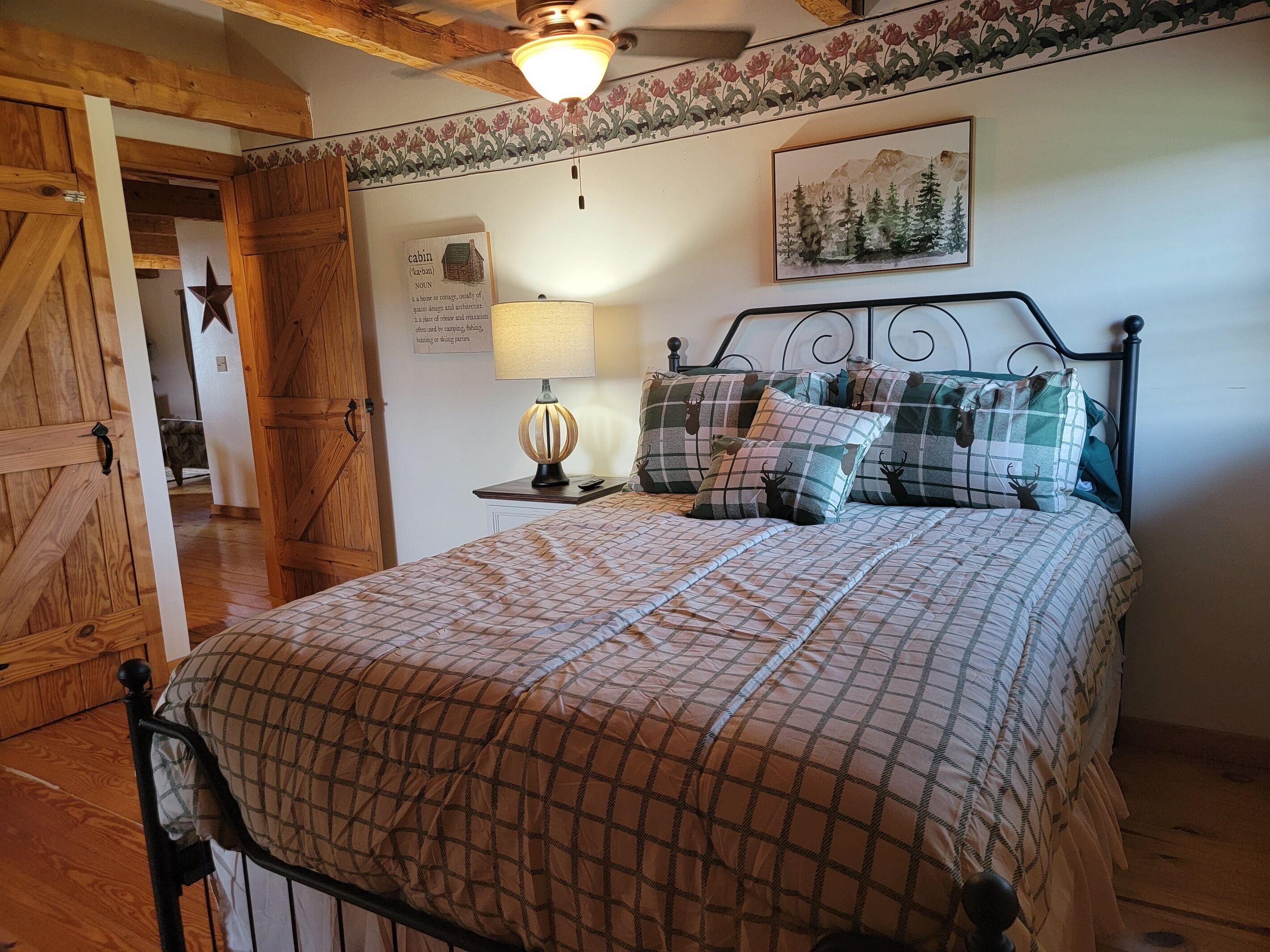 A bedroom with a wooden bed frame, a checkered bedspread, a nightstand with a lamp, and a wall-mounted painting.