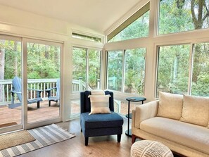 A room with a large glass door leading to an outdoor deck, a blue armchair, a beige sofa, and a small round table.